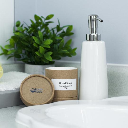 Waterless unscented soap in cardboard container with lid off, on the countertop with acrylic hand soap pump, sink, and plant beside the container.